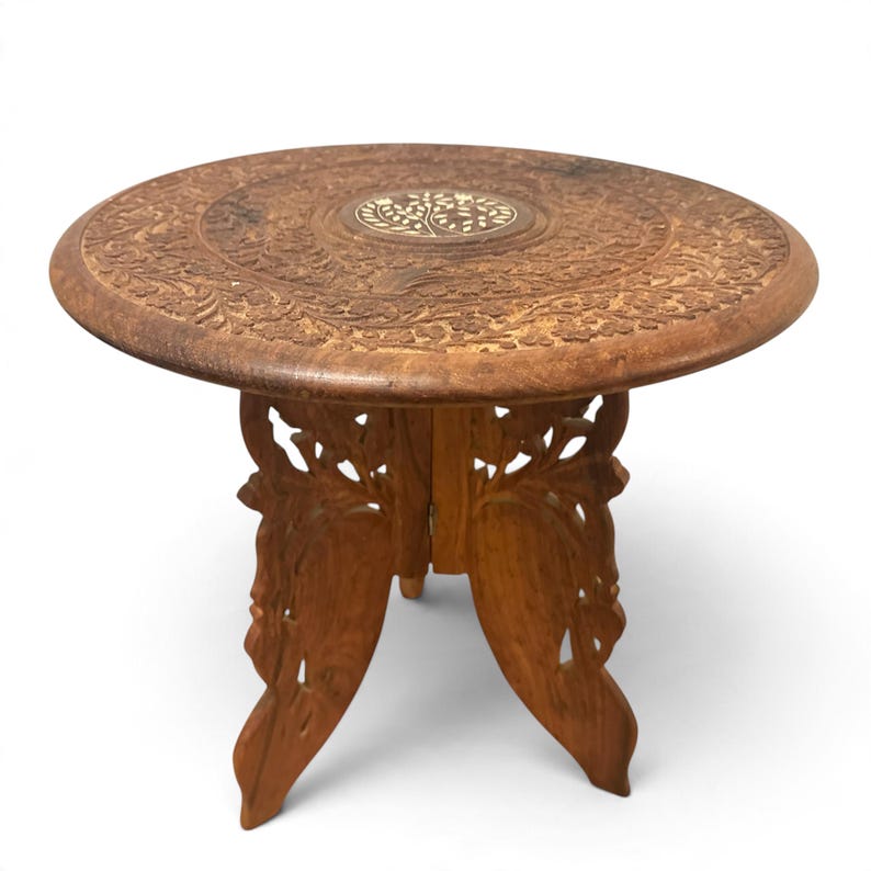 May include: A round wooden side table featuring intricate floral carvings across the surface and a central circular inlay depicting a tree. The base consists of two interlocking panels with openwork cutouts, finished in a warm brown tone.