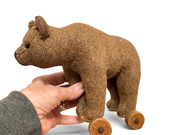 Puede incluir: Un juguete de oso de tela marrón con textura que se sostiene sobre cuatro pequeñas ruedas de madera. El oso tiene un hocico redondeado, orejas pequeñas y ojos oscuros. El cuerpo está cubierto de un material tejido grueso y cuatro ruedas.