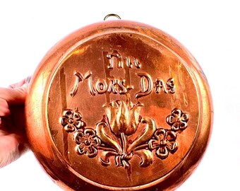May include: A round copper wall plaque featuring an embossed tulip and flower design at the bottom. The text "Tot Mors-Das" is centred in the upper portion. The metal has a polished finish with a small hanging loop at the top edge.