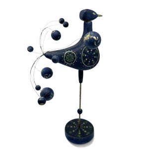 May include: A dark blue wooden bird figurine standing on a circular base. The bird features a long metal wire tail with attached blue spheres, and its body is decorated with green and white circular patterns. A thin metal rod supports the bird.