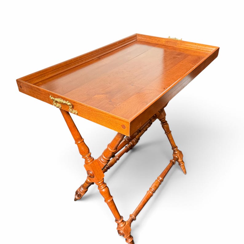 Puede incluir: Una mesa bandeja rectangular de madera con un acabado liso y pulido y un borde elevado. La bandeja cuenta con asas de metal dorado ornamentadas en los laterales y se apoya sobre una base plegable en forma de X con patas torneadas.
