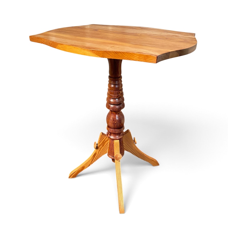 May include: A wooden side table featuring a smooth, irregularly shaped tabletop with a natural grain pattern. The table is supported by a central, vertically turned pedestal column that connects to a three-legged base structure.