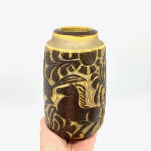 Peut inclure: Un vase en c&eacute;ramique marron et beige avec un motif sculpt&eacute; textur&eacute;. Le vase a une forme cylindrique avec un bord l&eacute;g&egrave;rement &eacute;vas&eacute;. Le motif sculpt&eacute; comprend des motifs floraux et abstraits. Le vase est tenu par une main.