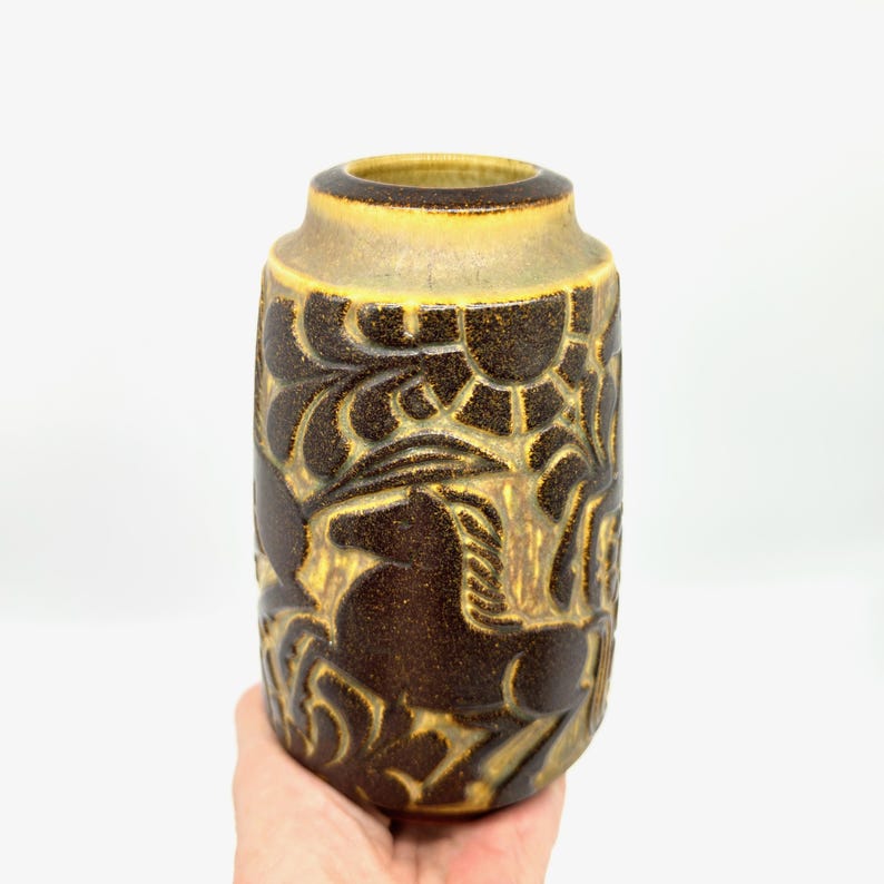 Peut inclure: Un vase en c&eacute;ramique marron et beige avec un motif sculpt&eacute; textur&eacute;. Le vase a une forme cylindrique avec un bord l&eacute;g&egrave;rement &eacute;vas&eacute;. Le motif sculpt&eacute; comprend des motifs floraux et abstraits. Le vase est tenu par une main.