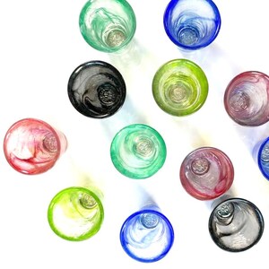 Puede incluir: Vista a&eacute;rea de una colecci&oacute;n de vasos de vidrio coloridos y arremolinados. Los vasos son en tonos de verde, azul, rosa, negro y amarillo. Los vasos est&aacute;n dispuestos sobre una superficie blanca, con sombras proyectadas debajo.