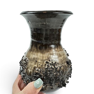 May include: A ceramic vase with a flared rim and a bulbous body featuring a dark brown and cream gradient glaze. The lower portion of the vase is covered in a textured, craggy, dark brown volcanic-style glaze that creates a rough, uneven surface.