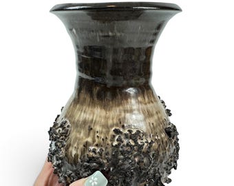 May include: A ceramic vase with a flared rim and a bulbous body featuring a dark brown and cream gradient glaze. The lower portion of the vase is covered in a textured, craggy, dark brown volcanic-style glaze that creates a rough, uneven surface.