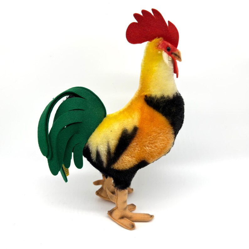 May include: A plush rooster figurine with a red comb, yellow head, and orange and black body. The rooster has a green tail and tan feet, standing on a white surface. This decorative item is a charming addition to any home decor.