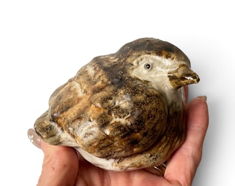 May include: A small ceramic bird figurine featuring a mottled brown and cream glaze with a glossy finish. The bird is depicted in a resting position with its head turned slightly to the side, showcasing detailed textures on the wings and body.