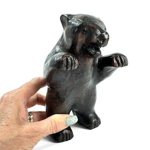 May include: A dark brown bear figurine standing on its hind legs with its front paws raised. The bear has a smooth, glossy finish, visible ears, a snout, and an open mouth. The sculpture is crafted with a textured surface and deep brown tones.