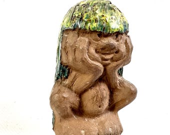 May include: A brown clay figurine of a seated creature with its hands pressed against its face. The figure has a textured, glossy green glaze covering the top of its head, resembling hair, and a smooth, matte brown finish on the body.