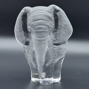 May include: A clear glass elephant sculpture. The elephant is shown from the front, with detailed texture on its skin, trunk, and ears. The sculpture is set against a dark background, highlighting its form.