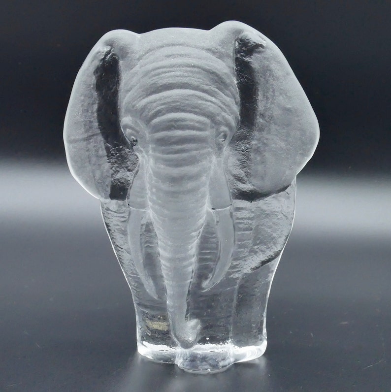 May include: A clear glass elephant sculpture. The elephant is shown from the front, with detailed texture on its skin, trunk, and ears. The sculpture is set against a dark background, highlighting its form.