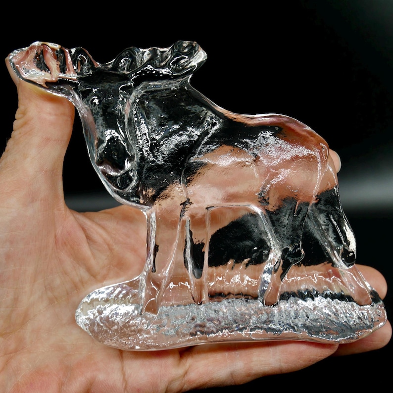 May include: A clear glass moose figurine with detailed antlers and body features. The moose is standing on a base, and the glass has a reflective quality. The figurine is held in a hand.