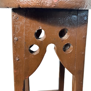 May include: A brown painted wooden stool featuring a circular top and four legs connected by a lower support. The upper section of each leg displays a cutout design with four circular holes and a central arch, secured with visible metal nails.