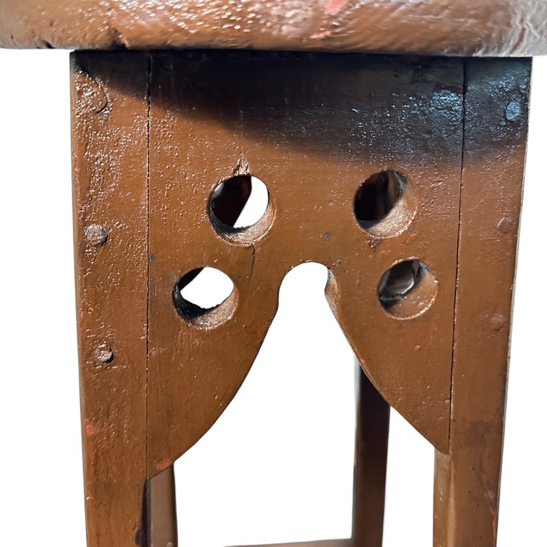 May include: A brown painted wooden stool featuring a circular top and four legs connected by a lower support. The upper section of each leg displays a cutout design with four circular holes and a central arch, secured with visible metal nails.