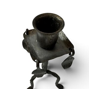 May include: A dark gray metal candle holder featuring a square base with rounded corners and a central cylindrical cup. The structure is supported by three twisted metal legs, with small decorative metal tabs hanging from the square base.
