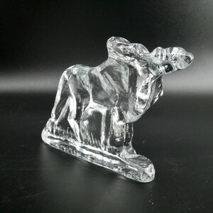 May include: A clear glass sculpture of a moose, with detailed features and a textured surface. The moose is depicted in a standing position, with its antlers visible. The sculpture is on a rectangular base.