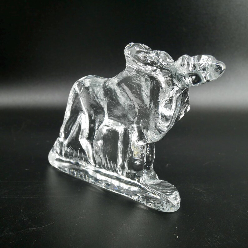 May include: A clear glass sculpture of a moose, with detailed features and a textured surface. The moose is depicted in a standing position, with its antlers visible. The sculpture is on a rectangular base.