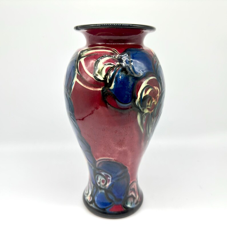 May include: A decorative ceramic vase with a glossy finish. The vase features a deep red base color with blue and white floral designs. The rim and base of the vase are black.