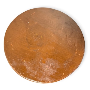 May include: A circular wooden board with a smooth, medium-brown finish. The surface shows signs of wear, including light scratches, minor scuffs, and small areas where the finish has faded, revealing a lighter wood tone underneath.