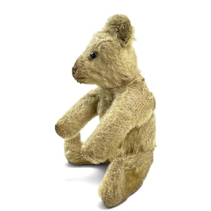May include: A vintage-style teddy bear crafted from light beige, textured mohair fabric. The bear is depicted in a seated position, featuring a prominent snout, small rounded ears, and dark, round eyes. The fur appears worn and coarse in texture.