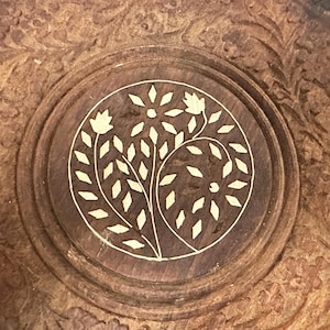 May include: A circular brown wooden object featuring an intricate carved border and a central inlay design. The inlay depicts a stylized floral motif with light-colored, diamond-shaped petals and leaves arranged within a defined circle.