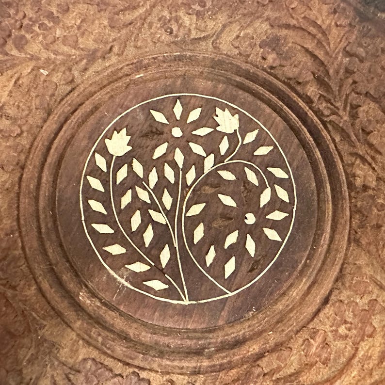 May include: A circular brown wooden object featuring an intricate carved border and a central inlay design. The inlay depicts a stylized floral motif with light-colored, diamond-shaped petals and leaves arranged within a defined circle.