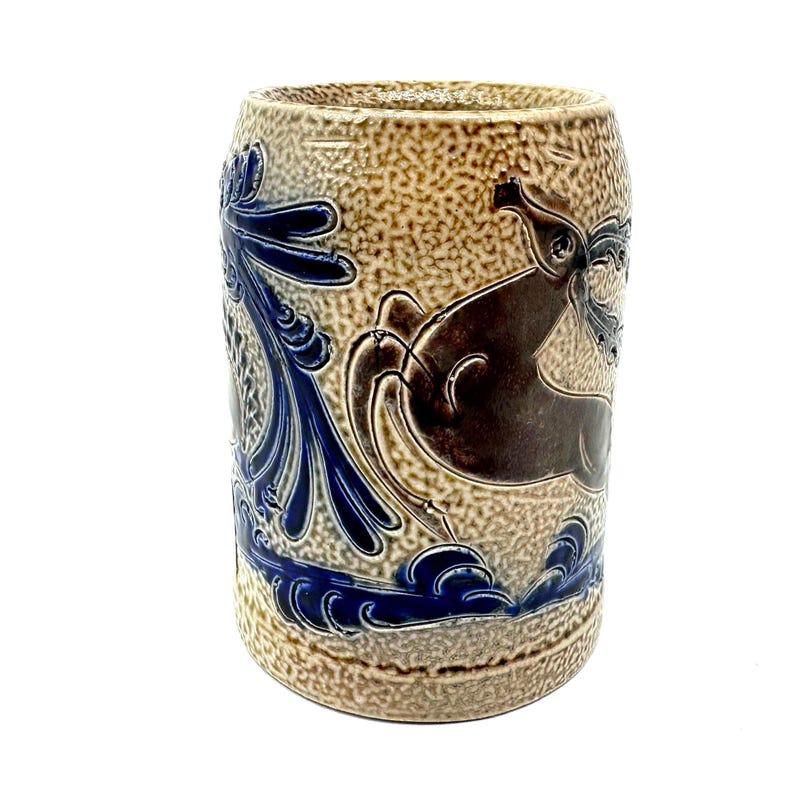 Puede incluir: Una jarra cil&iacute;ndrica de cer&aacute;mica con un acabado de esmalte de sal beige texturizado. El exterior est&aacute; decorado con patrones florales azul oscuro en relieve y una figura de ciervo marr&oacute;n que salta sobre la superficie, sobre un fondo punteado.