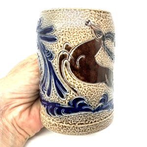 Puede incluir: Una jarra de cerveza cil&iacute;ndrica de cer&aacute;mica con un acabado beige texturizado de esmalte salino. El recipiente presenta un relieve de un ciervo marr&oacute;n saltando entre follaje y olas azules estilizadas. La base y los laterales son azul cobalto.