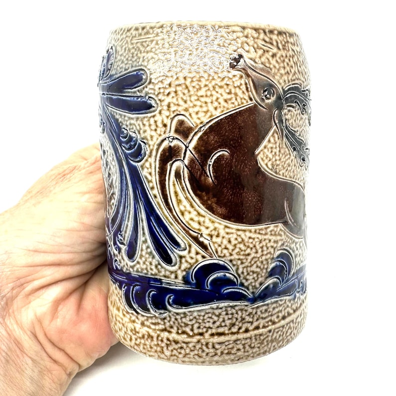 Puede incluir: Una jarra de cerveza cil&iacute;ndrica de cer&aacute;mica con un acabado beige texturizado de esmalte salino. El recipiente presenta un relieve de un ciervo marr&oacute;n saltando entre follaje y olas azules estilizadas. La base y los laterales son azul cobalto.