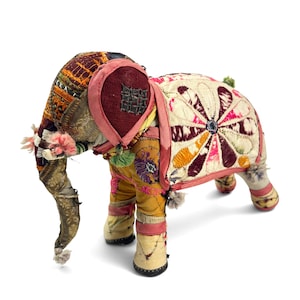 May include: A stuffed elephant figurine crafted from various patches of fabric. The body features a cream-colored back panel with a large, dark pink and orange floral-like embroidered design. The head and legs are covered in multicolored, textured textile scraps with intricate stitching and small decorative tassels.