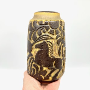 Peut inclure: Un vase en c&eacute;ramique marron avec une surface textur&eacute;e et une forme cylindrique. Le vase pr&eacute;sente un motif en relief de chevaux, de soleils et de feuillage. Le bord sup&eacute;rieur est d'une nuance de marron plus claire.
