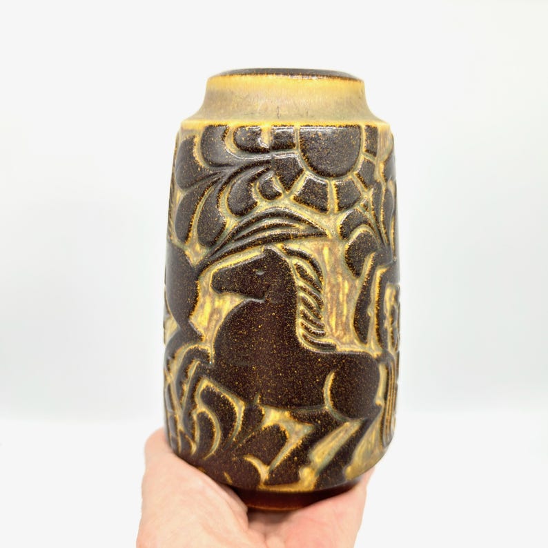 Peut inclure: Un vase en c&eacute;ramique marron avec une surface textur&eacute;e et une forme cylindrique. Le vase pr&eacute;sente un motif en relief de chevaux, de soleils et de feuillage. Le bord sup&eacute;rieur est d'une nuance de marron plus claire.