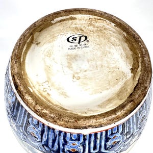Puede incluir: La parte inferior de un recipiente de cer&aacute;mica con un logotipo circular con las letras SP, el texto MADE IN CHINA y caracteres chinos. La base tiene un borde marr&oacute;n sin esmaltar, mientras que el cuerpo muestra un dise&ntilde;o azul, blanco y naranja.