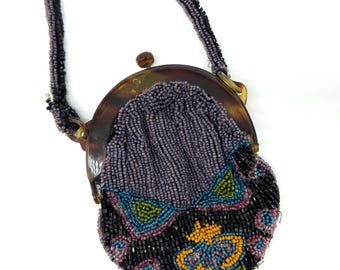 May include: A small round beaded coin purse featuring a dark brown tortoiseshell-style frame and a matching beaded strap. The purse is decorated with intricate patterns of purple, black, blue, green, and yellow glass beads in a floral design.