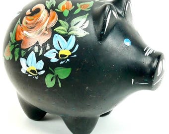 May include: A black ceramic piggy bank featuring hand-painted floral motifs in shades of orange, light blue, and green. The pig-shaped coin bank has a coin slot on the top, pointed ears, and simple white painted details on the snout and eyes.
