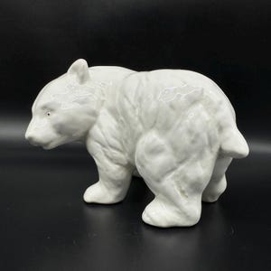 Könnte beinhalten: Eine weiße Keramik-Bärenfigur. Der Bär wird in einer Laufpose dargestellt, mit detaillierter Fellstruktur und glänzendem Finish. Die Figur ist ein dekorativer Artikel, geeignet zur Präsentation.