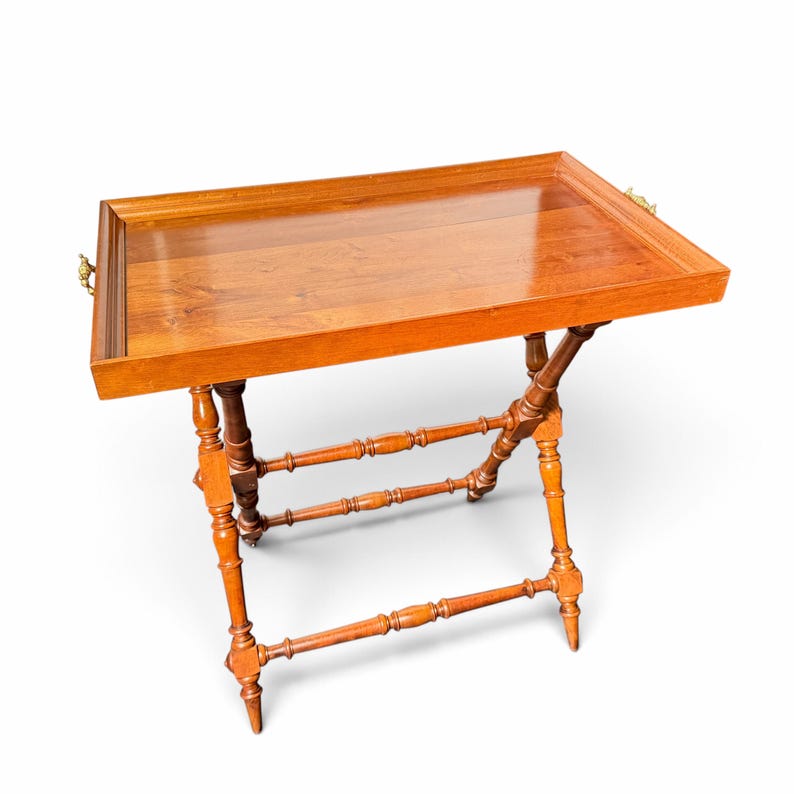 Puede incluir: Una mesa bandeja de madera rectangular con un acabado marr&oacute;n pulido. La bandeja presenta bordes elevados y asas de metal dorado en los laterales. Se apoya sobre un soporte plegable con cuatro patas torneadas y travesa&ntilde;os.