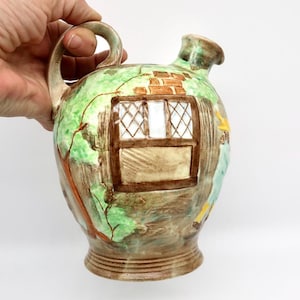 May include: A ceramic pitcher featuring a hand-painted scene of a cottage exterior with a brown window frame, white lattice panes, and a brick chimney. The vessel has a rounded body, a curved handle, and a green and brown glazed finish.