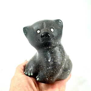Op de afbeelding: Zwarte keramische berenfiguur met witte ogen en een glimlachende uitdrukking. De beer heeft een gespikkelde textuur en zit met zijn poten naar voren. Het beeldje is een decoratief item.