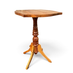 May include: A wooden pedestal table featuring a triangular top with rounded corners and a visible wood grain pattern. The central support column is turned with decorative bulbous sections, resting on a three-legged base with curved feet.