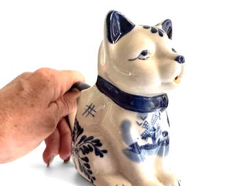 Op de afbeelding: Een keramische kan in de vorm van een kat met een glanzend crèmekleurig glazuur, handgeschilderde blauwe bloemmotieven, een afbeelding van een windmolen op de zijkant en een donkerblauwe halsband. De kan heeft een handvat en een schenktuit.
