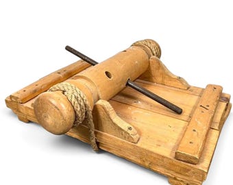 May include: A light brown wooden device featuring a central cylindrical roller with a thick rope wrapped around one end. A dark metal rod passes through the center of the cylinder, which is mounted on a flat wooden base with side supports.