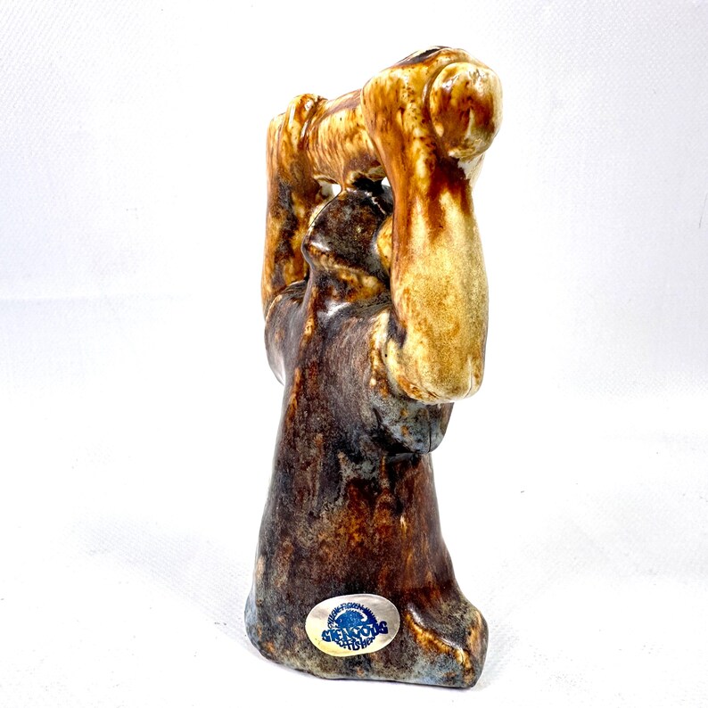 May include: A ceramic figurine featuring a figure with arms raised above the head, finished in a mottled brown and cream glaze. The base of the sculpture displays a small oval sticker with the blue text Glencoe Pottery visible.