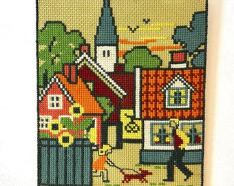 May include: A rectangular needlepoint tapestry featuring a village scene with red and orange houses, a tall church steeple, and green trees. A person walks a small red dog on a leash near a fence with yellow sunflowers against a tan background.
