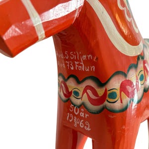 Puede incluir: Una figura de caballo de madera de color naranja brillante con motivos florales pintados a mano en blanco, rojo y azul cerceta. El lateral muestra el texto blanco Avd.5 Siljan, Avd 73 Falun, 50 &aring;r, 19 20 62, con detalles de arn&eacute;s pintados en blanco.