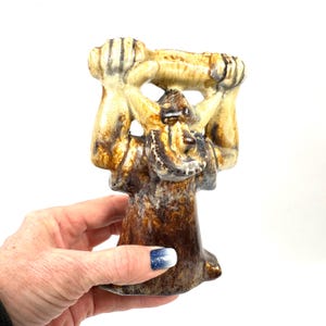 May include: A ceramic figurine in shades of brown, tan, and cream. The sculpture depicts a stylized figure holding a horizontal object above its head. The figurine has a textured surface and a unique, artistic design. The base is a darker brown.