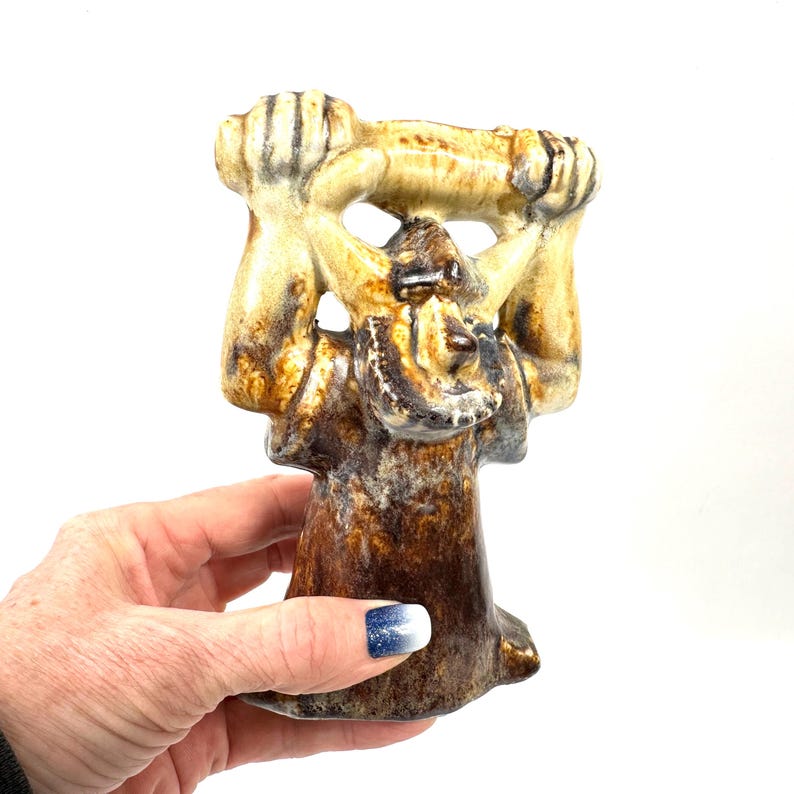 May include: A ceramic figurine in shades of brown, tan, and cream. The sculpture depicts a stylized figure holding a horizontal object above its head. The figurine has a textured surface and a unique, artistic design. The base is a darker brown.