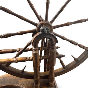 May include: A dark brown wooden spinning wheel featuring a large circular wheel with multiple spokes, a central hub, and a sturdy base. The structure includes turned wooden supports and a horizontal spindle assembly with detailed carved accents.